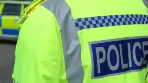 BBC A close up of a police officer wearing a high visibility yellow jacket with police written on it.
