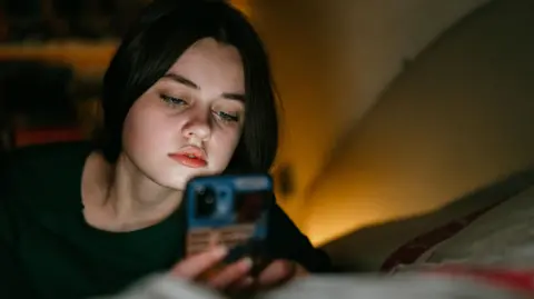 Portrait of a teenage girl looking at a mobile phone. She is laying on her bed. She has dark hair and a green jumper on and there is a light in the right of the photo.