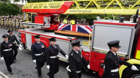 PA Media A turntable ladder carrying the coffin of Firefighter Martyn Sadler passes Bicester Fire Station, Oxfordshire, ahead of his funeral. The coffin in draped in the Union Flag and the vehicle is flacked by firefighters in full dress unifor