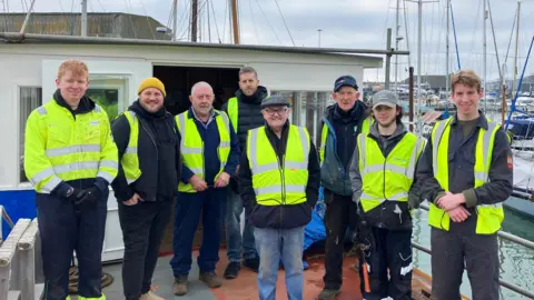 Jon Wright/BBC Eight people stand in a line on a boat deck all wearing high-visibility jackets. They are all looking at the camera and smiling. 