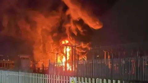 London Fire Brigade Flames shoot into the nighty sky from the sub-station, which is surrounded by two sets of metal fencing, one with a CCTV warning sign on it.