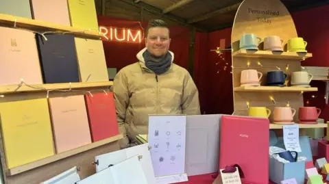 A man with short brown hair stands behind a stall selling colourful personalised notebooks and mugs.