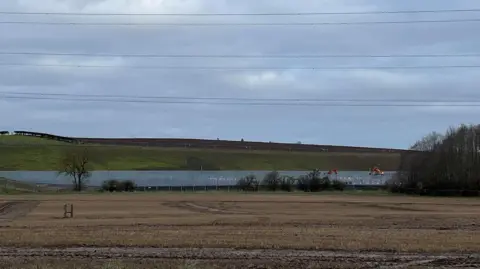 A newly fenced off area where construction work is taking place, with a crop field in the foreground and grassy hillside to the rear