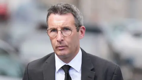 PA  Muir is outside. He's wearing a black suit with a white short and a black tie. 