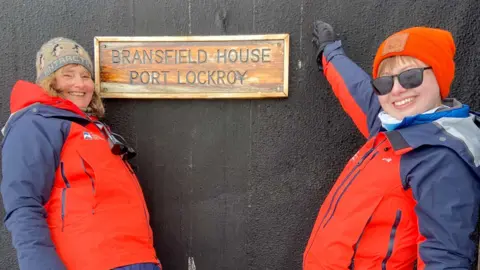 UKAHT/Lisa Ford On the right is Aoife McKenna wearing  hat and glasses and a red coat and pointing at a sign saying "Bransfield House Port Lockroy". On the left is another team member wearing a red coat.