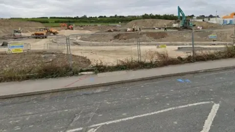 Google A road in the front of the image, with a building site in the distance. Diggers and trucks are working at the construction site.