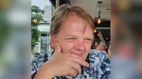 A man with mid length brown hair parted in the middle, looks away from the camera. His hand is up to his face and he is resting his cheek on his thumb. he is wearing a white and black patterned shirt.