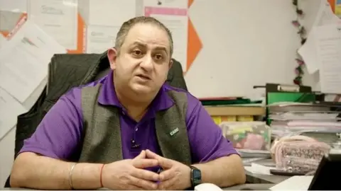 Raj Sehgal is sitting at his desk with his hands touching. He has short brown hair, and is clean shaven. He is wearing a purple polo shirt and a grey waistcoat. Behind him are files of paperwork.