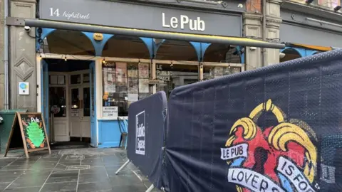 BBC The outside of Le Pub, which has a grey sign with white writing. There is also  a sign outside that says "Le Pub is for lovers"  