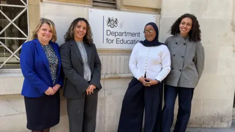 A group of four women, including the campaign founder and Carole Gould, standing beside an outdoor sign which says 'Department for Education'.