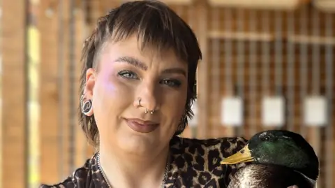 A woman with short brown hair is looking into the camera, smiling. She has stretchers in her ears, and two piercings in her nose. She is wearing a leopard print top. She is holding a duck to the right of the image. It has a green head, a yellow beak, and a brown feathered body. 