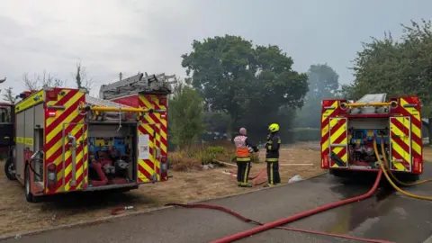 Two fire vehicles are at the scene of the emergency. Two officers are stood talking to each other in between the vehicles. A hose can be seen coming from the vehicle. One of the people talking is wearing a green helmet.
