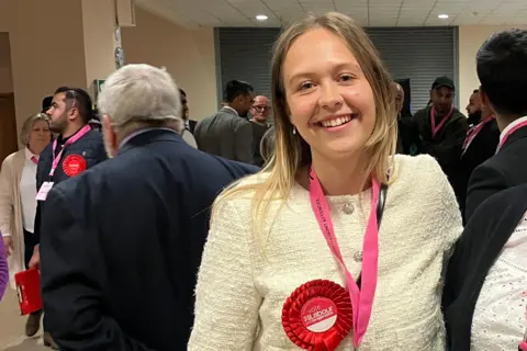 Shariqua Ahmed/BBC Daisy Creedon-Blakemore, a young woman with blonde hair who is wearing a white jacket with a red Labour rosette. She is standing inside a hall or foyer area with lots of people smartly dressed behind her - mostly men in dark-coloured suits.