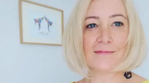 A person with a short blonde bob haircut is shown in a close-up indoors, wearing a yellow top and with a small dark tattoo visible on the side of the neck. In the background, there is a light-coloured wall with a framed picture of two dogs and a wooden cross-like decoration mounted on it.