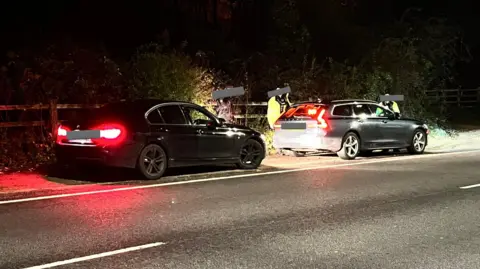 A BMW and an unmarked police car pulled over at the side of the A1 road. Three people are standing near the vehicles and their faces have been blocked out