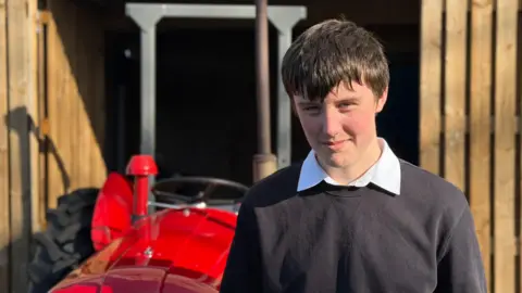 A boy in a school jumper and shirt stands in front of the red tractor. He has straight dark hair and rosy red cheeks. He is squinting as there is a low sun in his eyes.