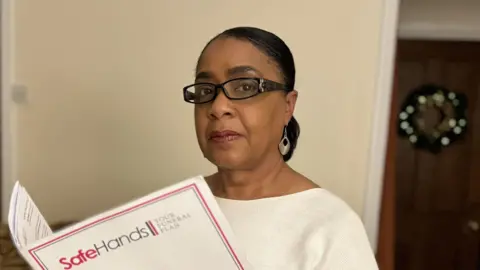 BBC A woman wearing a white jumper and glasses is holding a folder with the Safe Hands branding.