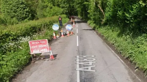 Google Temporary lights are on the road, next to traffic cones. Nearer to the camera is a sign saying "when red light shows wait here". There are trees to the left and right of the road.