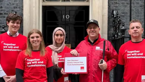 Ray Clements Ray poses in the middle with two women and two men outside the black door of 10 Downing Street. They smile as they pose in red tops saying "Together we will end homelessness" and Ray and a lady int he middle hold a sign calling on Keir Starmer to "build a future free from homelessness".