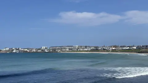 A scenic view overlooking the west strand in Portrush with apartments and homes far off in the distant. It is a sunny day with a blue sky and a blue sea with waves crashing.