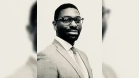 A sepia image of Damo Awobajo, a man wearing glasses, a suit jacket plus a shirt and tie.