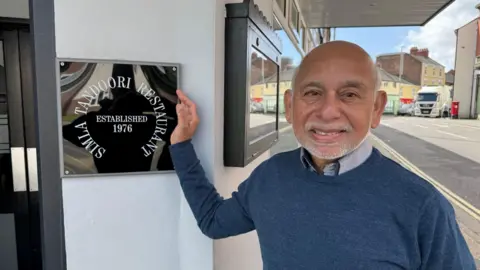 This image shows a man standing outside a building entrance, placing one hand on a wall-mounted plaque. The plaque reads “Simla Tandoori Restaurant – Established 1976.” The person is wearing a blue sweater and collared shirt and is positioned beside a doorway. In the background, a street scene is visible with buildings, parked vehicles and road markings, suggesting a town centre setting.