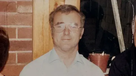Julie Westwood An older man with short grey hair who is wearing aviator glasses with a clear frame. He is smiling and pictured in front of a wall that is exposed brick. 