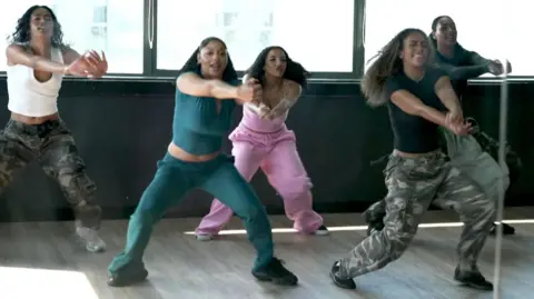 Five members of the dance troupe dance a routine with their arms stretched in front of them and their legs stood apart. They are wearing leisurewear in various colours, including teal, pink and black