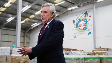 PA Media A man with grey hair and who is wearing a black suit jacket with a white shirt and red tie, in front of a warehouse full of boxes. 