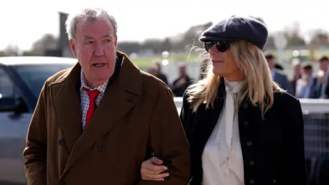 PA Media Jeremy Clarkson strolls arm in arm with his partner Lisa Hogan. Clarkson wears a brown overcoat, checked shirt and red ties. Hogan wears a black jacket, white silk shirt and a baker boy hat.