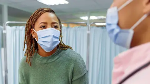 Getty Images A woman wearing casual clothing sits on a hospital bed and speaks with her doctor. Both are wearing blue face masks.