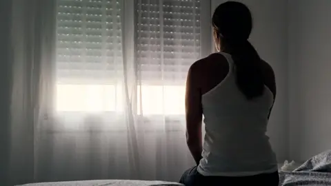 A woman wearing a white vest and her hair in a ponytail sitting on a bed facing a window.