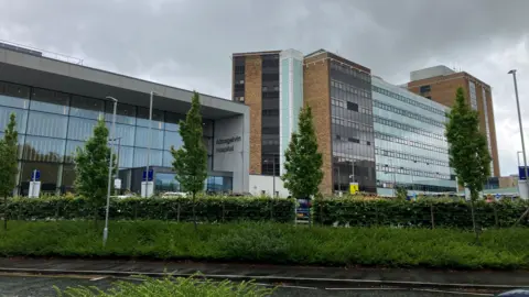 A wide shot of Altnagelvin Hospital on a cloudy day. It is a tall, multi-story building with brown brick, glass and other materials featured in its front elevation. The main entrance features a tall, glass-fronted atrium attached to the main tower block. Hedges and trees line the road outside the building.