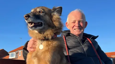 Cola sat on a bench with John Dalley who has his arm around the dog and a red lead draped round his black padded jacket