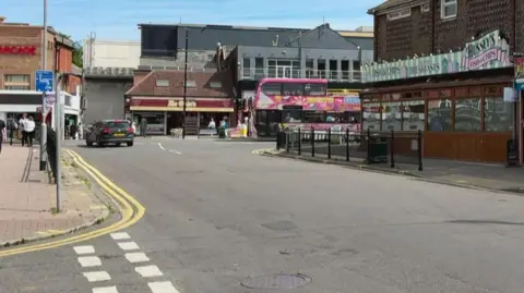 A view of Drummond road.  There is fish and chip shop in the building to the right, and a pink double decker bus crosses the t junction at the bottom of the road.