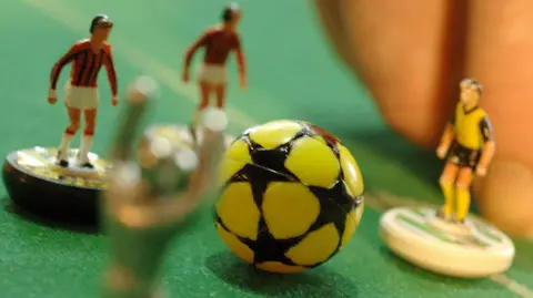 Getty Images A close up of a Subbuteo set. There are three miniature plastic football player figurines on a grass like pitch. Someone is flicking one of the players with their finger in front of a yellow and black football.