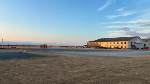 A small cream building airport. It is surrounded by green grass and fields. It also has car parks all around.