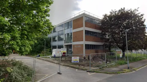 A Google street view screenshot of a brick building in an industrial site with trees on either side. The site is fenced off.