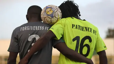  one from Forgiveness FC (L) - in a black shirt, the other from  Patience FC (R) - in a green shirt. They pose a football between their heads after playing a peace football match. Archive shot 2021.
