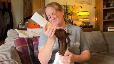Tanya Bishop wearing a grey top bottle feeding a brown and white baby goat