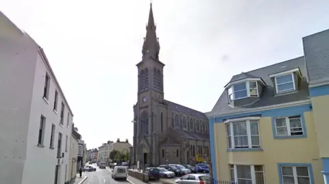 Google Street View image of St Thomas Church in Jersey. It has a tall spire. A yellow and blue building is next to it and several other buildings are on the opposite side of the road. Several vehicles are parked in a car park next to the church. It is a cloudy day.