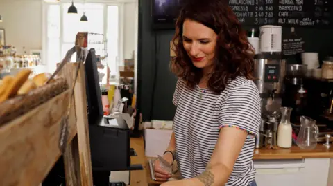 woman with shoulder-length brown hair in stripey t-shirt cleaning tables in a cafe