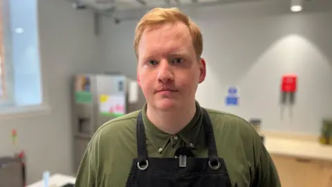 Peter Gilchrist, with ginger hair and wearing a green T-shirt and black apron, is standing in an industrial kitchen.