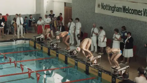 Mark Williams Four disabled swimmers are photographed just as they are about to jump into a pool at the start of a race. Mark is on the starting block that is furthest away from the camera and is wearing a white swimming cap with his amputated leg lifted as he gets ready to launch.