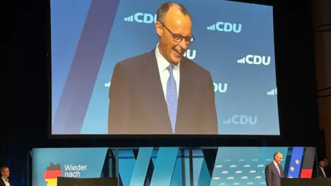 BBC Friedrich Merz grins on a stage with a big screen as a backdrop and a light-blue backing with the slogan in German "Back in front"