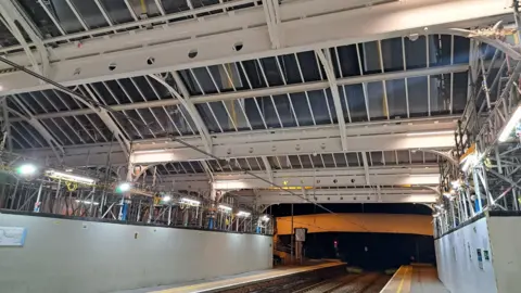 Work on the canopy above the tracks has almost been completed. It has new glass panes and the frames have been painted white. The station is well lit but empty of passengers. 