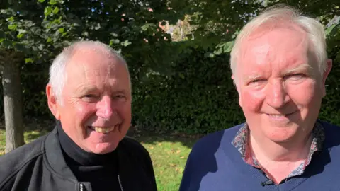 BBC Nick Owen and Ian Phillipson sitting together and smiling. Nick wears a black jacket and a black turtleneck sweater. Ian wears a blue sweater over a colorful shirt.