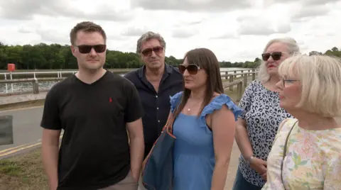 BBC A group of men and women stand in front of a road by the side of Earlswood Lakes.
