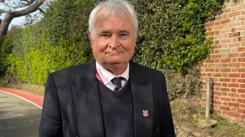 Mark Labey has white hair and is wearing a black suit, jumper, white shirt and tie. He has a pin badge on his left lapel. Behind him there is a country lane with trees, hedges and part of the road painted red with a white outline, defining the walking path.