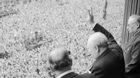 Winston Churchill on a balcony holding his hand up in a V for victory sign for a large crowd